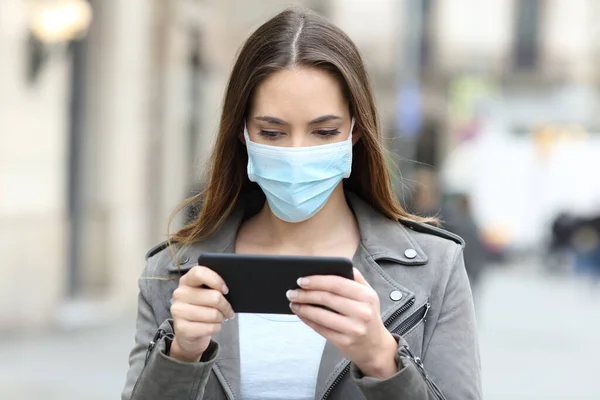 Serious woman with protective mask avoiding contagion watching video online on smart phone in the street