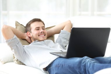 Mutlu adam dizüstü bilgisayarda film seyrediyor. Evdeki kanepede yatıyor.