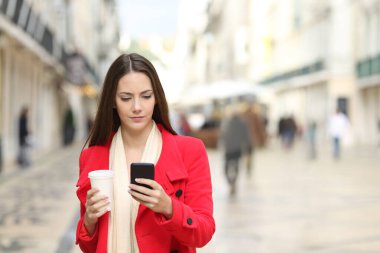 Kırmızı paltolu ciddi bir kadının kışın sokakta yürürken elinde hazır kahveyle akıllı telefon kullanması ön planda.