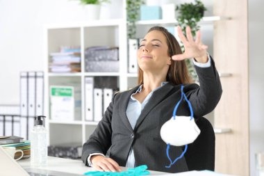 Satisfied executive woman breathing fresh air throwing protective mask on a desk at the office clipart