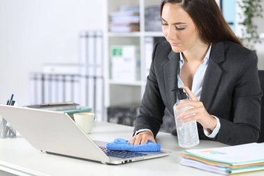 Yönetici kadın ofisteki bir masanın üstünde dezenfektan ve bezle dizüstü bilgisayarı dezenfekte ediyor.