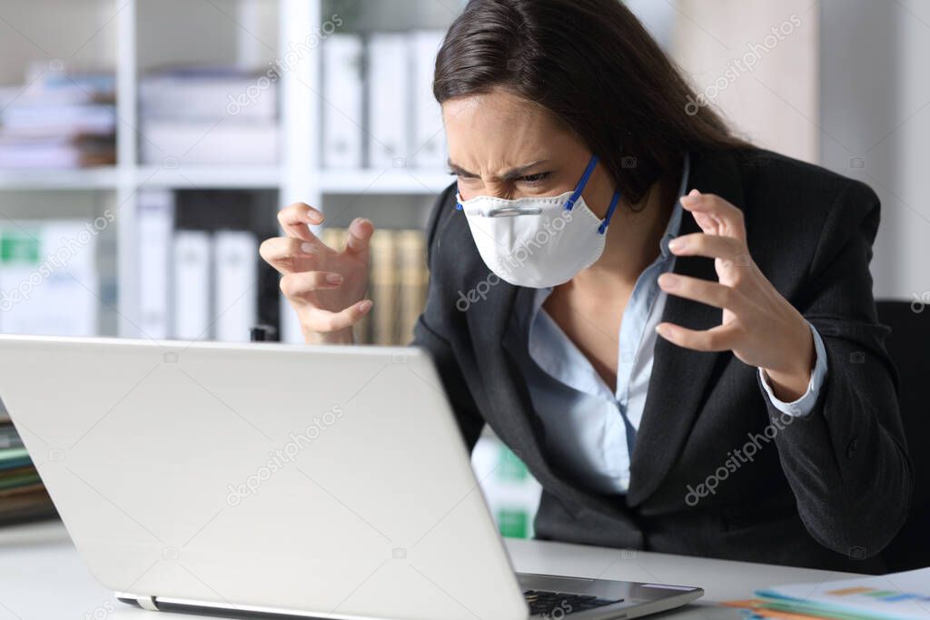 Angry executive woman reading bad news on laptop avoiding covid-19 with mask on a desk at the office