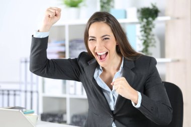 Heyecanlı yönetici kadın ofisteki masasında oturan kameraya bakarak iyi haberi kutluyor.