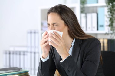 Executive woman sneezing using tissue paper covering mouth sitting on a desk at office clipart