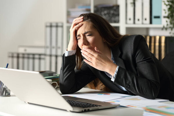 Уставшая исполнительная женщина зевает, работая допоздна, сидя за столом ночью в офисе
