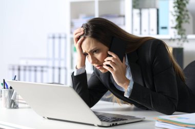 Üzgün yönetici kadın, ofisteki masasında oturmuş bilgisayarından akıllı telefon okuması yapıyor.
