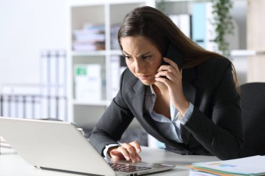 Suspicious executive woman calling on smart phone looking at laptop sitting on her desk at office clipart