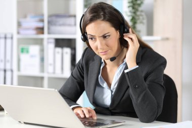 Suspicious telemarketer woman with headset looking at laptop sitting on a desk in the office clipart
