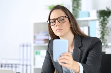 Executive woman wearing eyeglasses with eyesight problem trying to read on smart phone sitting on her desk at office clipart