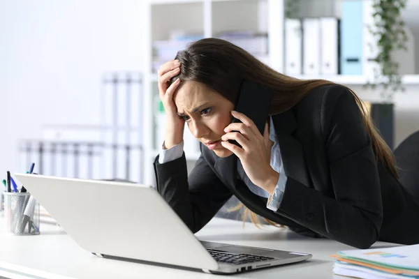 Sad executive woman calling on smart phone reading on laptop sitting on her desk at office