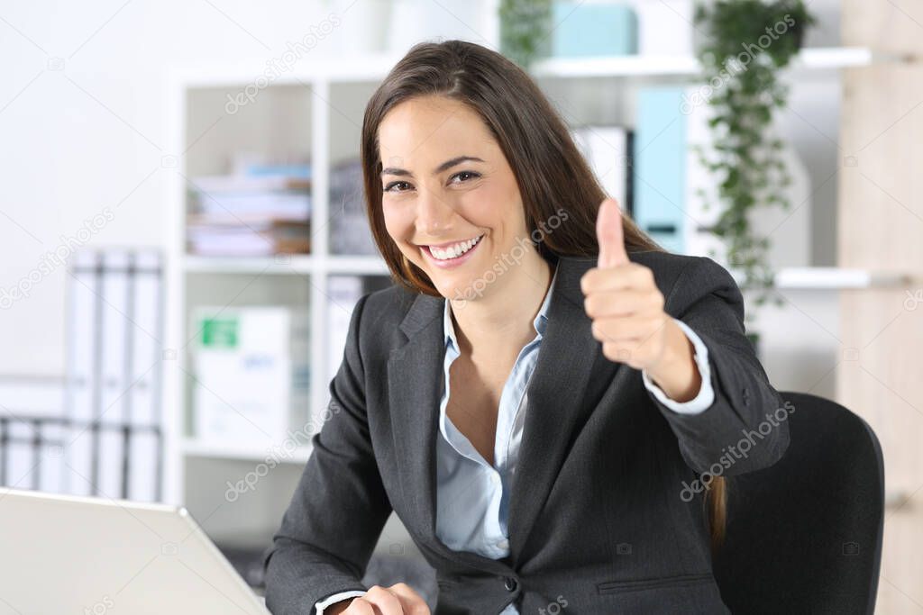 Happy executive woman gesturing thumbs up looking at camera sitting on a desk at the office