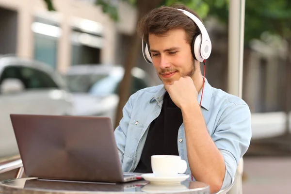 Man with headphones watches video on laptop sitting on a coffee shop terrace