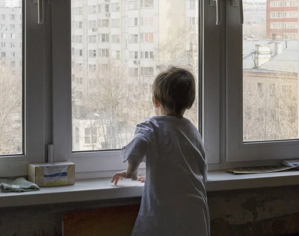 Mały chłopiec wygląda przez okno. Ulica jest jasna i słoneczna. Zimna pogoda — Zdjęcie stockowe