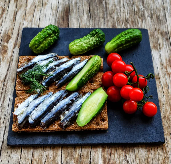 Ansjovis met verse komkommer en cherrytomaatjes op de zwarte rechthoekige plaat op de vintage houten tafel, traditioneel gepaard met wodka — Stockfoto