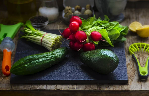 Cooking salad from fresh vegetables: cucumber, avocado, radishes, wild leek (ramson) arranged on the black tile, vintage wooden table with cutlery. Darkmood rustic style — Stock Photo, Image