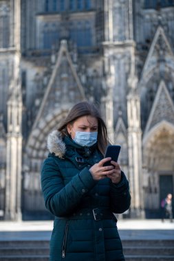 Koronavirüs (COVID-19) karantinasında koruyucu tıbbi maskeli genç bir kadın Almanya, Avrupa 'daki Köln Katedrali önünde akıllı telefon kullanıyor.