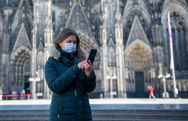 young woman with protective medical mask in quarantine of coronavirus (COVID-19) using smart phone in front of the Cologne Cathedral in Germany, Europe