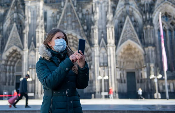 ドイツ ケルン大聖堂前のスマートフォンを使用したコロナウイルス Covid の隔離医療用マスクを持つ若い女性 ストック画像