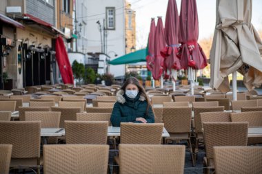 Almanya, Köln 'deki Coronavirus salgını sırasında boş kafeterya terasında oturan koruyucu maskeli ciddi bir genç kadın.