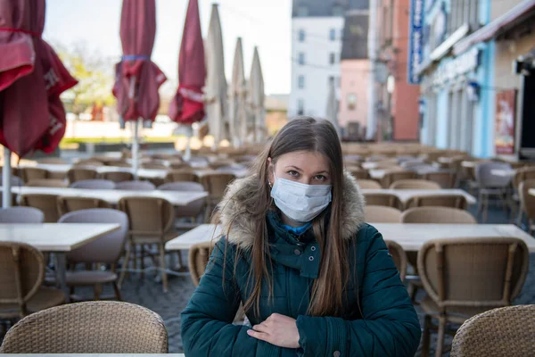 Serious young woman with protective face mask sitting outdoors at the empty cafe terrace  during the coronavirus outbreak in Cologne, Germany