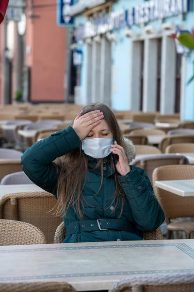 Serious young woman with protective face mask sitting outdoors at the empty cafe terrace and talking with somebody on her mobile phone during the coronavirus outbreak. having headache