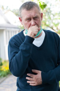  Güneşli bahar gününde arka bahçede ameliyat maskesiyle öksüren adam. Orta yaşlı Avrupalı adam