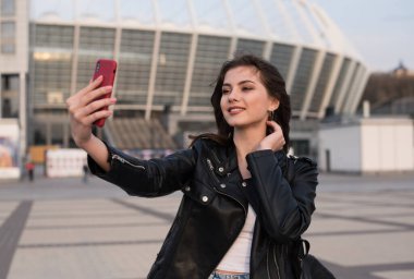 Güzel genç bir kadın şehirde selfie çekiyor. Gülümseyen Avrupalı bir kadın deri ceket giyiyor.
