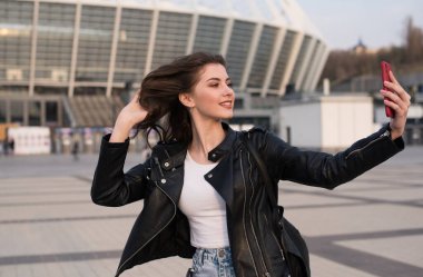 Gülümseyen genç kadın şehirde selfie çekiyor, Avrupalı kadın deri ceket giyiyor
