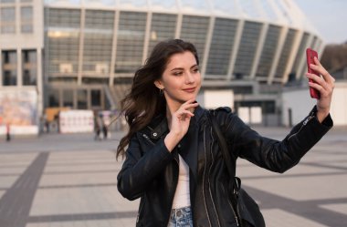 Gülümseyen genç kadın şehirde selfie çekiyor, Avrupalı kadın deri ceket giyiyor