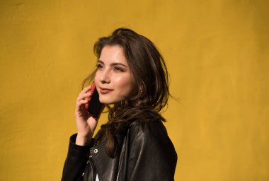 Deri ceketli güzel beyaz kız parlak turuncu arka planda telefonla konuşuyor. Genç mutlu kadın güneşli bir günde şehirde