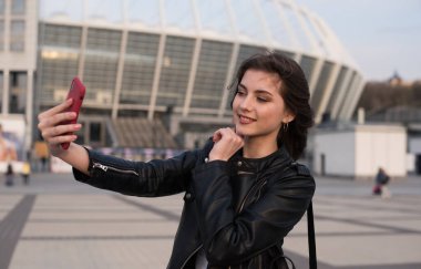 Gülümseyen genç kız şehirde selfie çekiyor, Avrupalı kadın deri ceket giyiyor