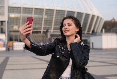 Gülümseyen genç beyaz kadın şehirde selfie çekiyor, Avrupalı kadın deri ceket giyiyor