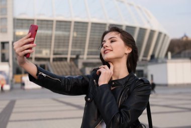 Gülümseyen genç kadın şehirde selfie çekiyor, Avrupalı kadın deri ceket giyiyor
