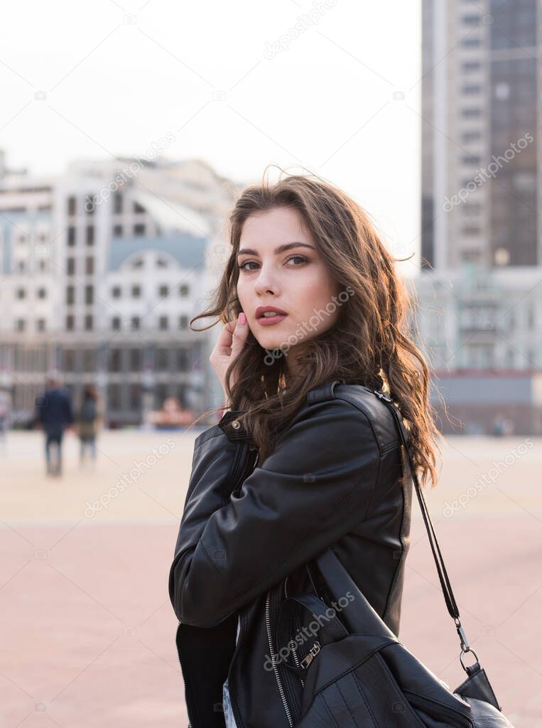 portrait of pretty brunette student girl in leather jacket on city background