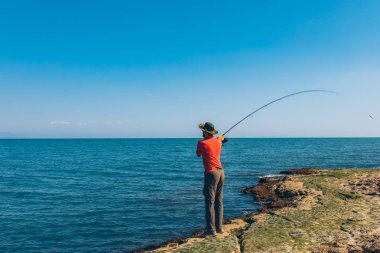 Balıkçı yaz mevsiminde denizde balık tutarken ayakta durup olta atıyor. Kayalıklarda ve deniz kıyısında bir balıkçı. Temiz mavi gökyüzü ve okyanusun altında balık tutmak