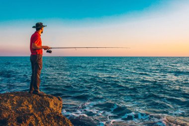 Balıkçı yaz mevsiminde denizde balık tutarken ayakta durup olta atıyor. Kayalıklarda ve deniz kıyısında bir balıkçı. Temiz mavi gökyüzü ve okyanusun altında balık tutmak
