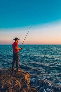 Balıkçı yaz mevsiminde denizde balık tutarken ayakta durup olta atıyor. Kayalıklarda ve deniz kıyısında bir balıkçı. Temiz mavi gökyüzü ve okyanusun altında balık tutmak
