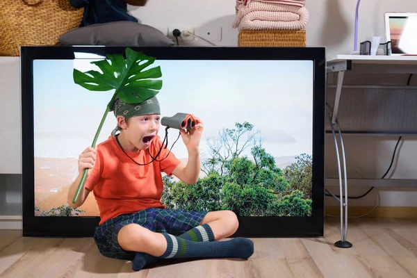 stock image Boy sitting and imitating scouting with field-glass at home near TV screen. Funny hiking, camping. Family time. Crazy activities. Coronavirus situation. Quarantine. Isolation from covid-19