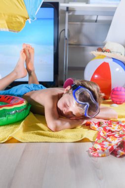 Çocuk karantina sırasında deniz aşırı tatilleri evdeki TV ekranının yanında taklit ediyor ve komik plaj aktivitelerinin tadını çıkarıyor. Coronavirus durumu. Evde kal. İzolasyon. Covid 19