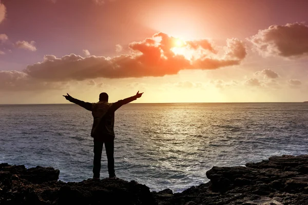 Uomo con le braccia distese sulle scogliere, Tramonto nuvoloso al mare — Foto Stock