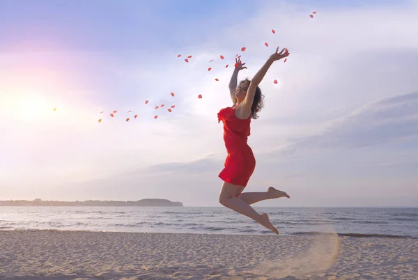 Άλμα όμορφη γυναίκα στο κόκκινο φόρεμα ρίψη αυξηθήκατε πέταλα στην παραλία. — Φωτογραφία Αρχείου