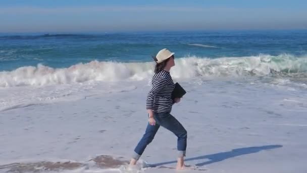 Mujer feliz corriendo y saltando en la playa con portátil en sombrero de paja — Vídeo de stock