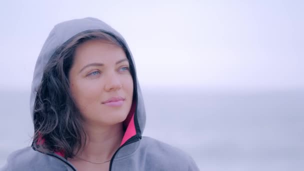 Mujer sonriente en impermeable en la playa gris lluviosa — Vídeos de Stock