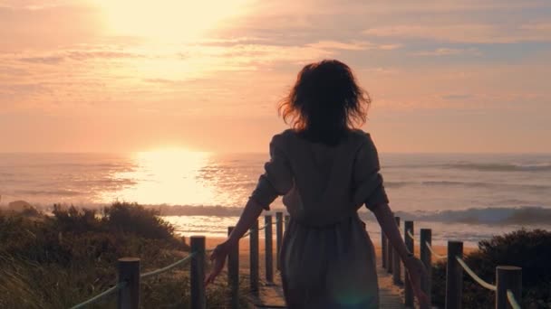 Mujer joven silueta en un puente de madera a pie de playa al atardecer. — Vídeos de Stock