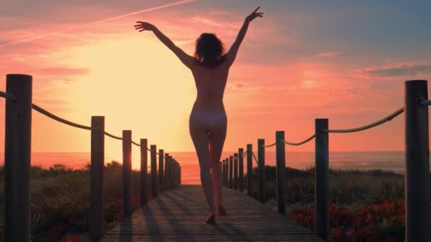 Mujer joven silueta en un puente de madera a pie de playa al atardecer. — Vídeo de stock