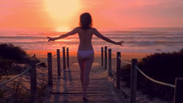 Jonge vrouw bikini in een houten voet brug op het strand bij zonsondergang — Stockvideo