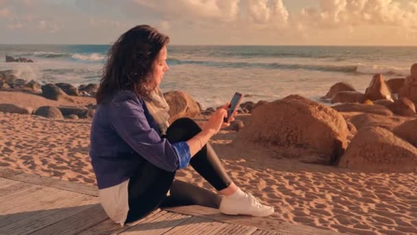 Mujer joven usando teléfono inteligente en la playa, turista tomando fotos de la puesta del sol — Vídeos de Stock