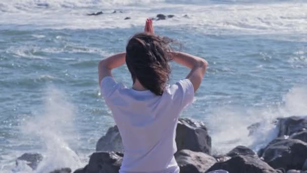 Женщина медитирует в позе лотоса на скалах у берега океана — стоковое видео