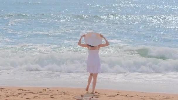 Mladá žena v bílých šatech a klobouku stojící na pláži — Stock video