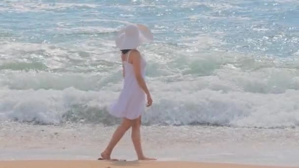 Ung kvinna bär en vit klänning och hatt promenader på en strand — Stockvideo